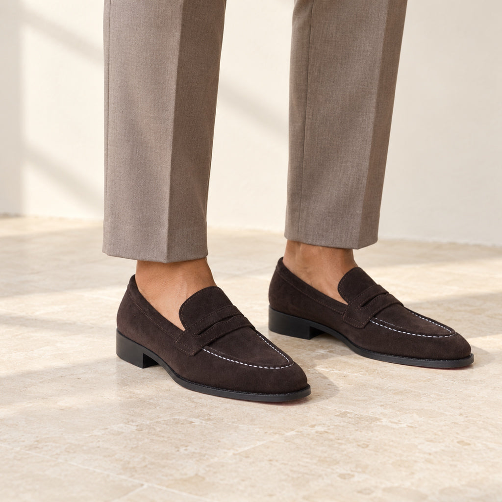 Expresso Brown Suede penny loafers worn with tailored trousers by a model with medium brown skin, cropped below the knees.